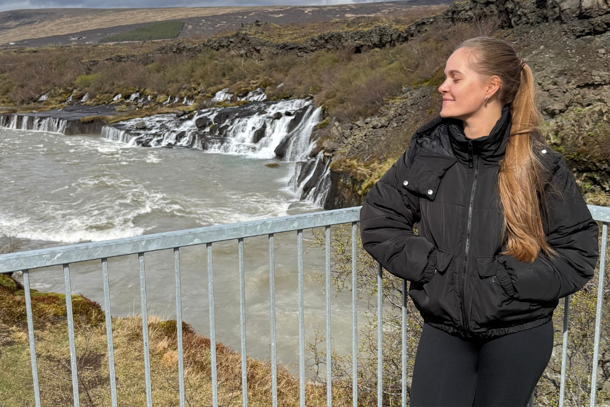 Anhelina from Arctic Adventures at a viewing platform above Hraunfossar waterfall, the wide lava-fed cascades and Hvítá river behind her.
