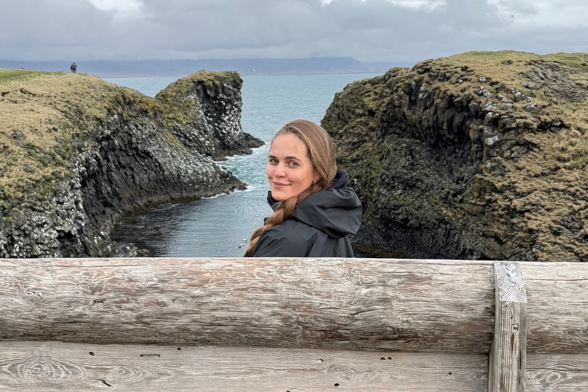 Anhelina looking back from a bench on the Arnarstapi coastal path, turquoise water and basalt cliffs behind her on the Snæfellsnes Peninsula.