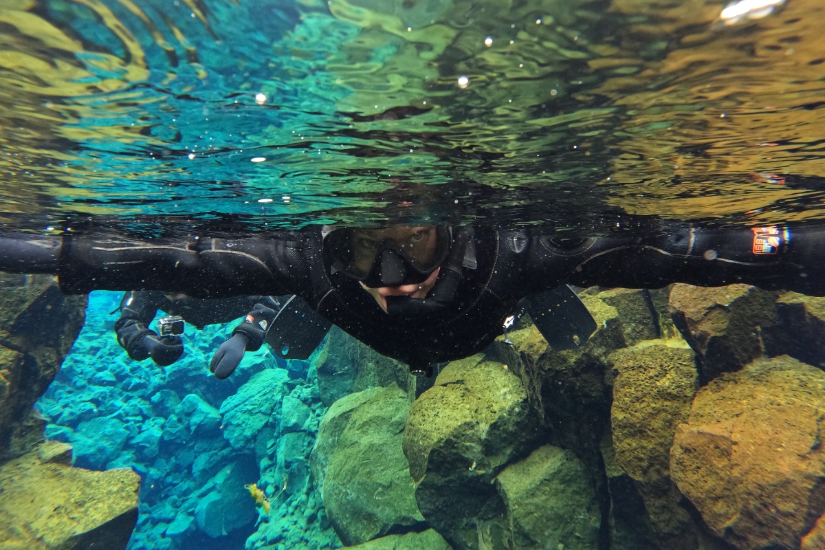Eva Sadler snorkeling underwater through the Silfra fissure, drysuit and mask on, between mossy rock walls.