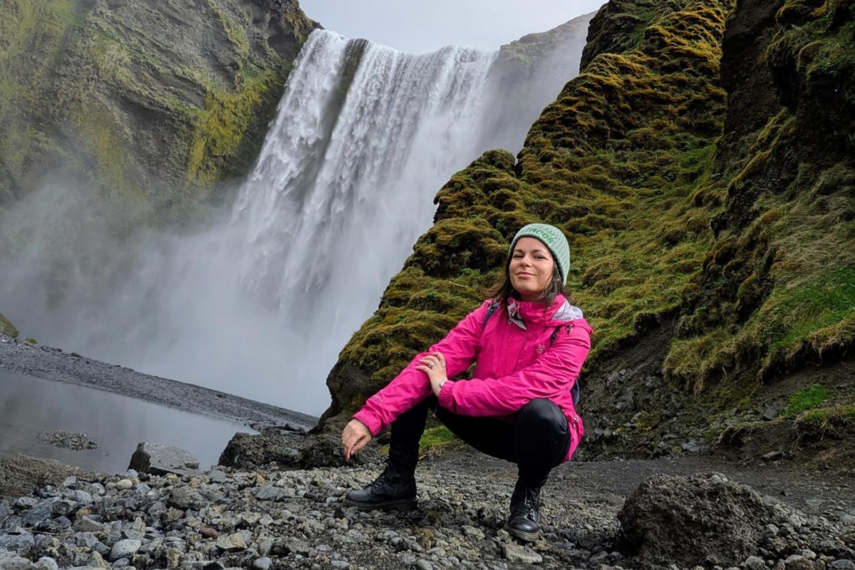 Eva crouching on the rocks in front of Skógafoss waterfall, mist rising behind her.