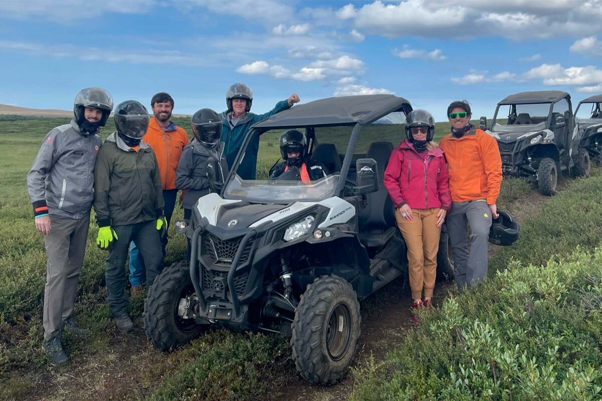 Group of riders in helmets and jackets beside an off-road buggy on green tundra in Iceland.
