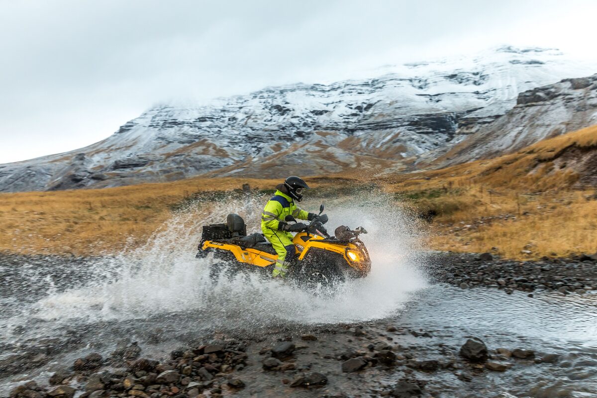 Rider in a yellow suit driving an ATV through a river with snow-capped mountains behind in Iceland.