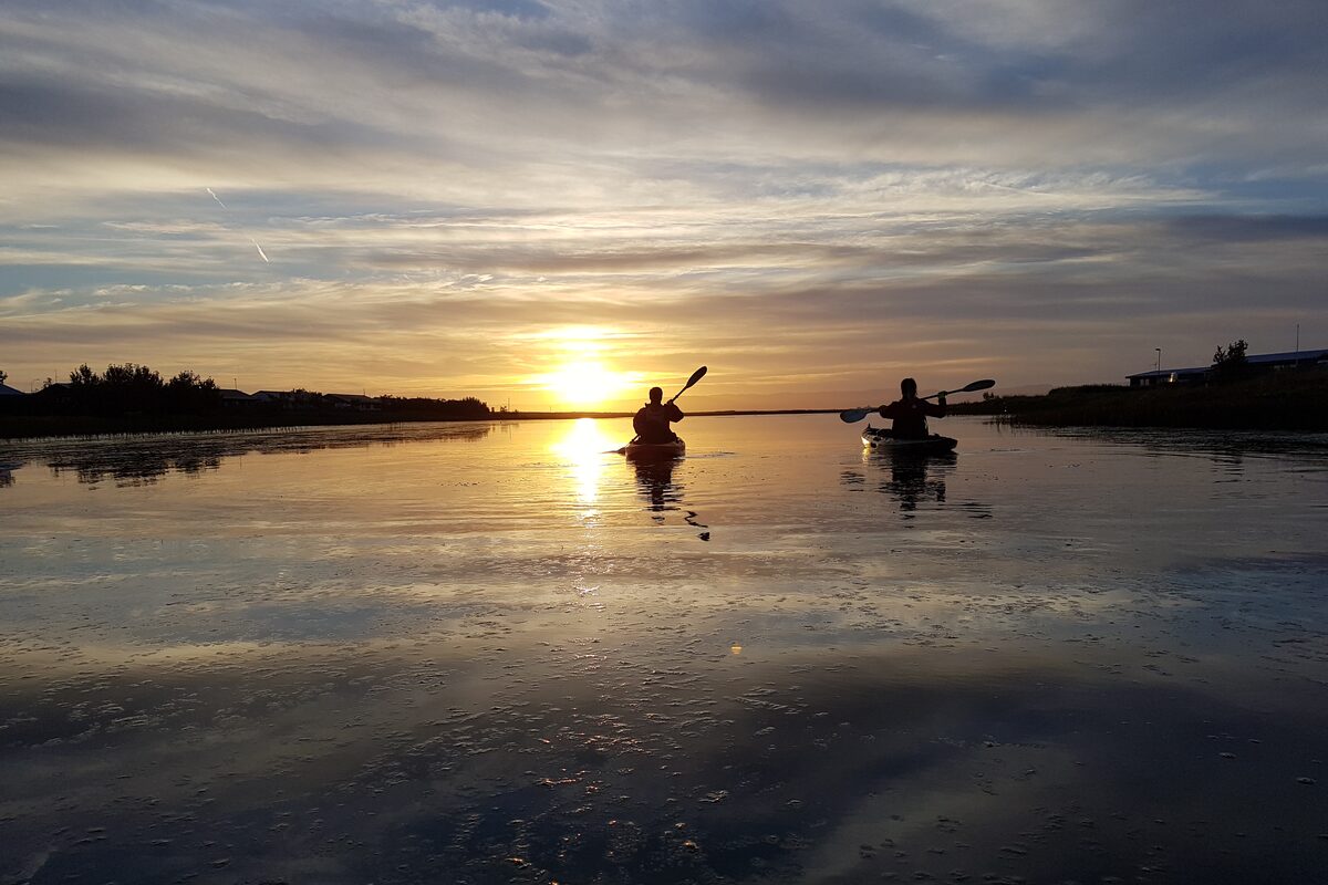 Two kayakers in silhouette paddle toward a low sun over mirror-calm water.
