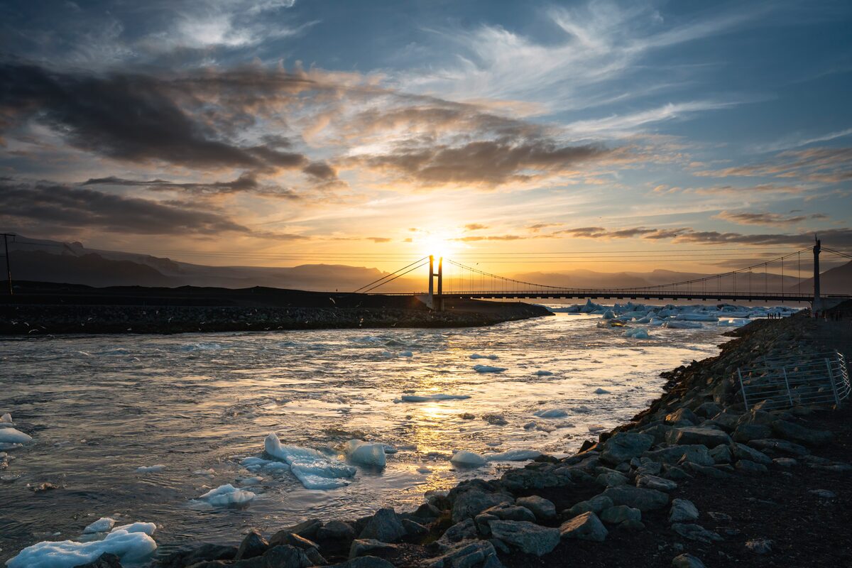 Golden sunset over water with floating ice and a distant bridge on an Icelandic shoreline.