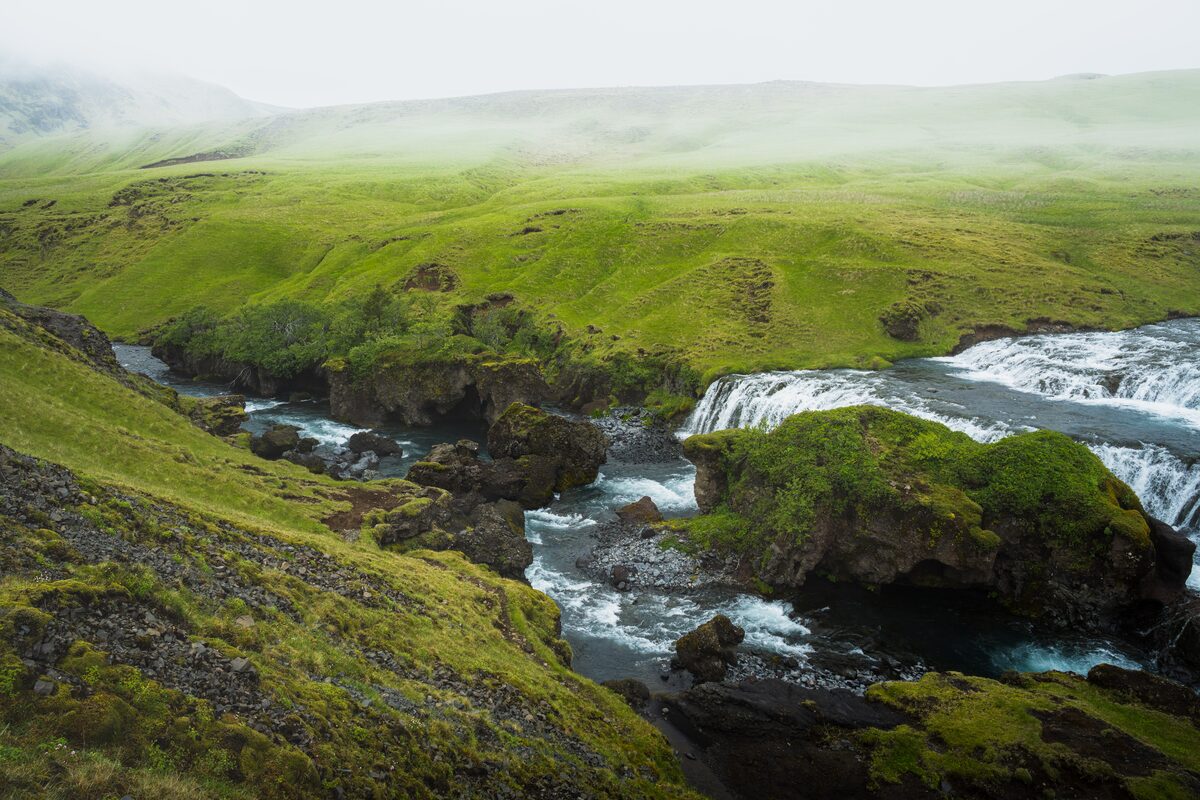 A river dropping over small waterfalls between mossy green hills under low mist in Iceland.