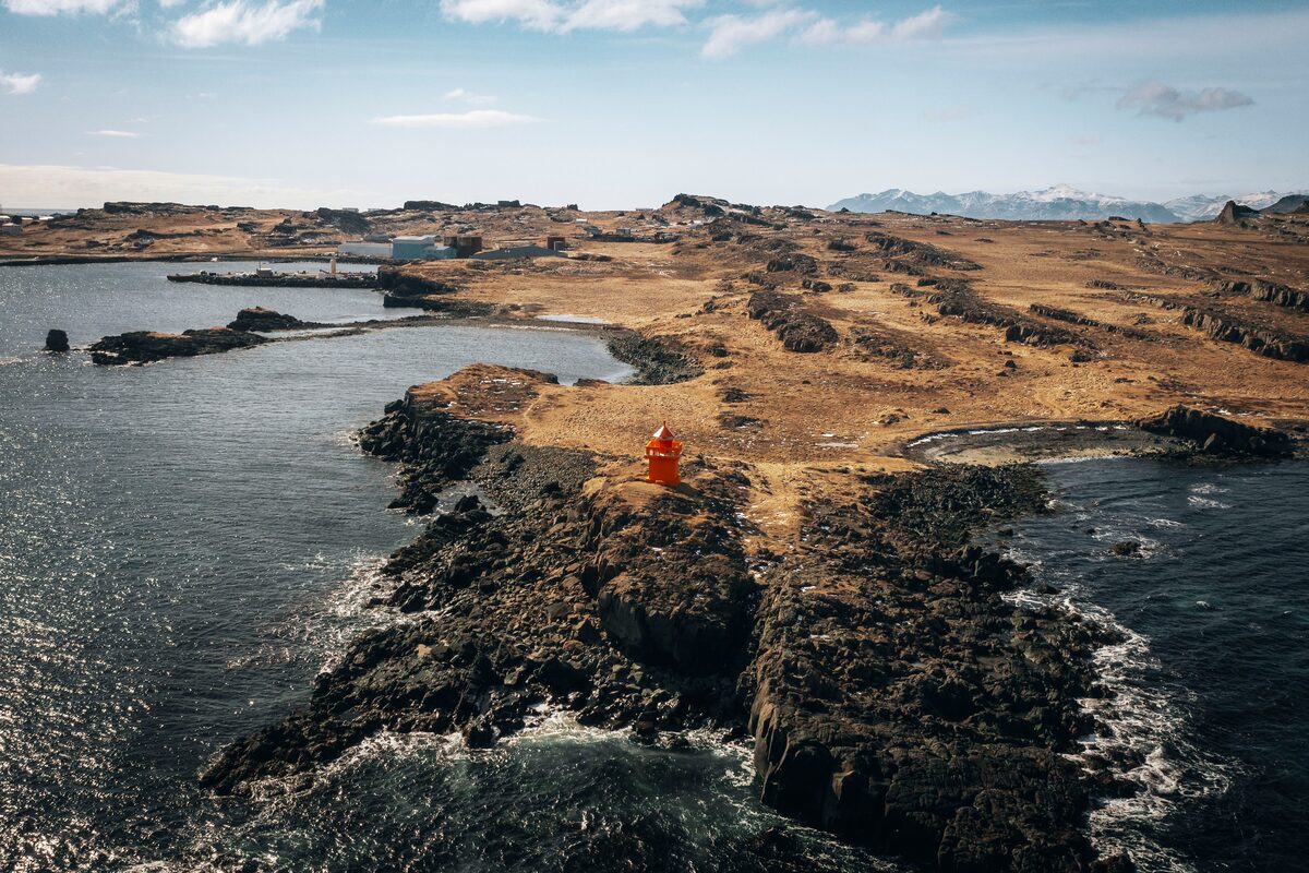 A red lighthouse on a rocky coastline in Iceland, with distant snow-topped mountains.