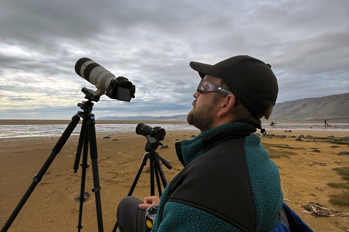 Photographer Gunnar Gaukur watches the sky beside camera tripods on a Westfjords black-sand beach