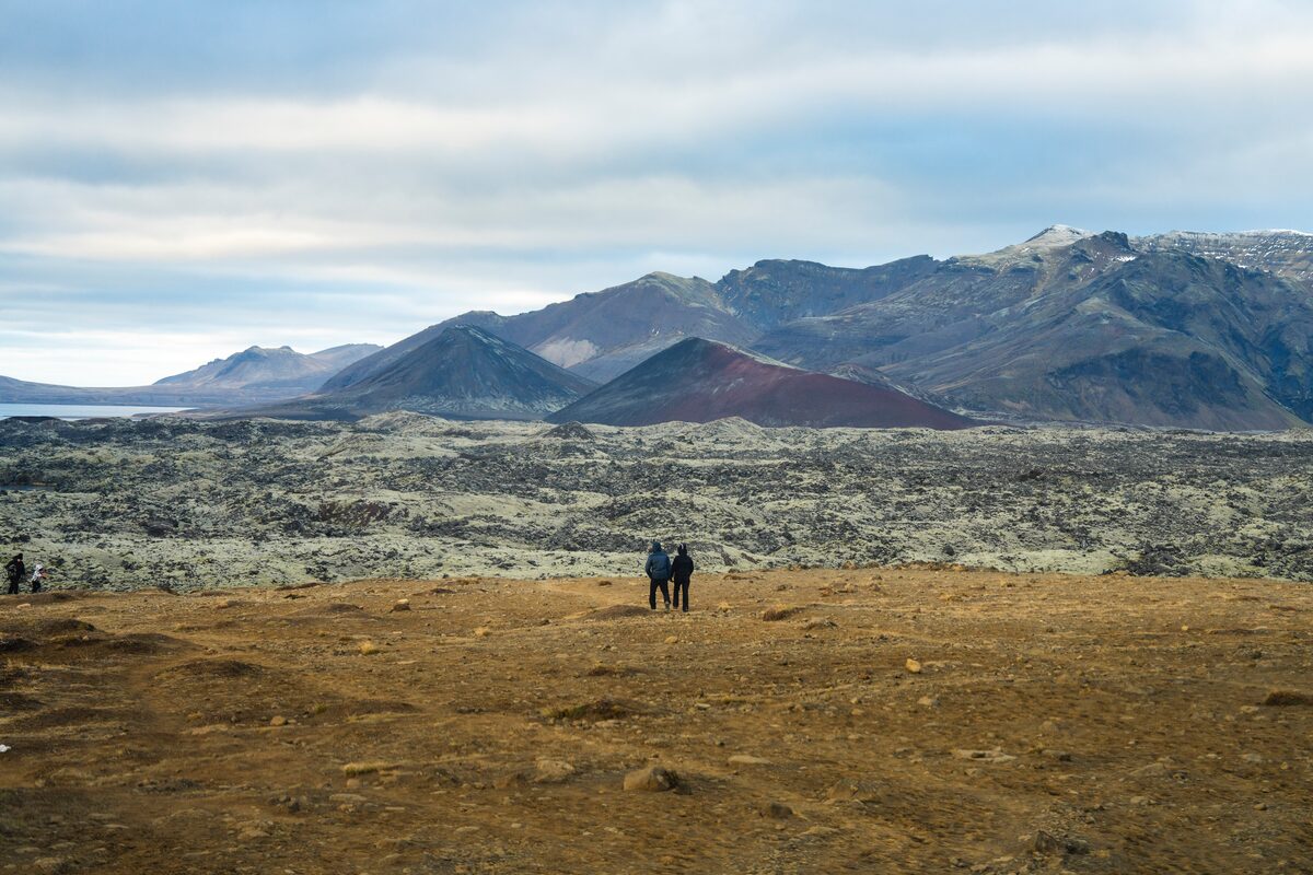 Two walkers on open ground before a mossy lava field on Snæfellsnes, red cone and mountains behind.