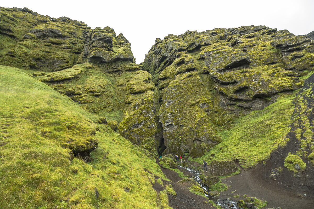 A narrow cleft in moss-covered cliffs with tiny figures at its entrance and a stream below.
