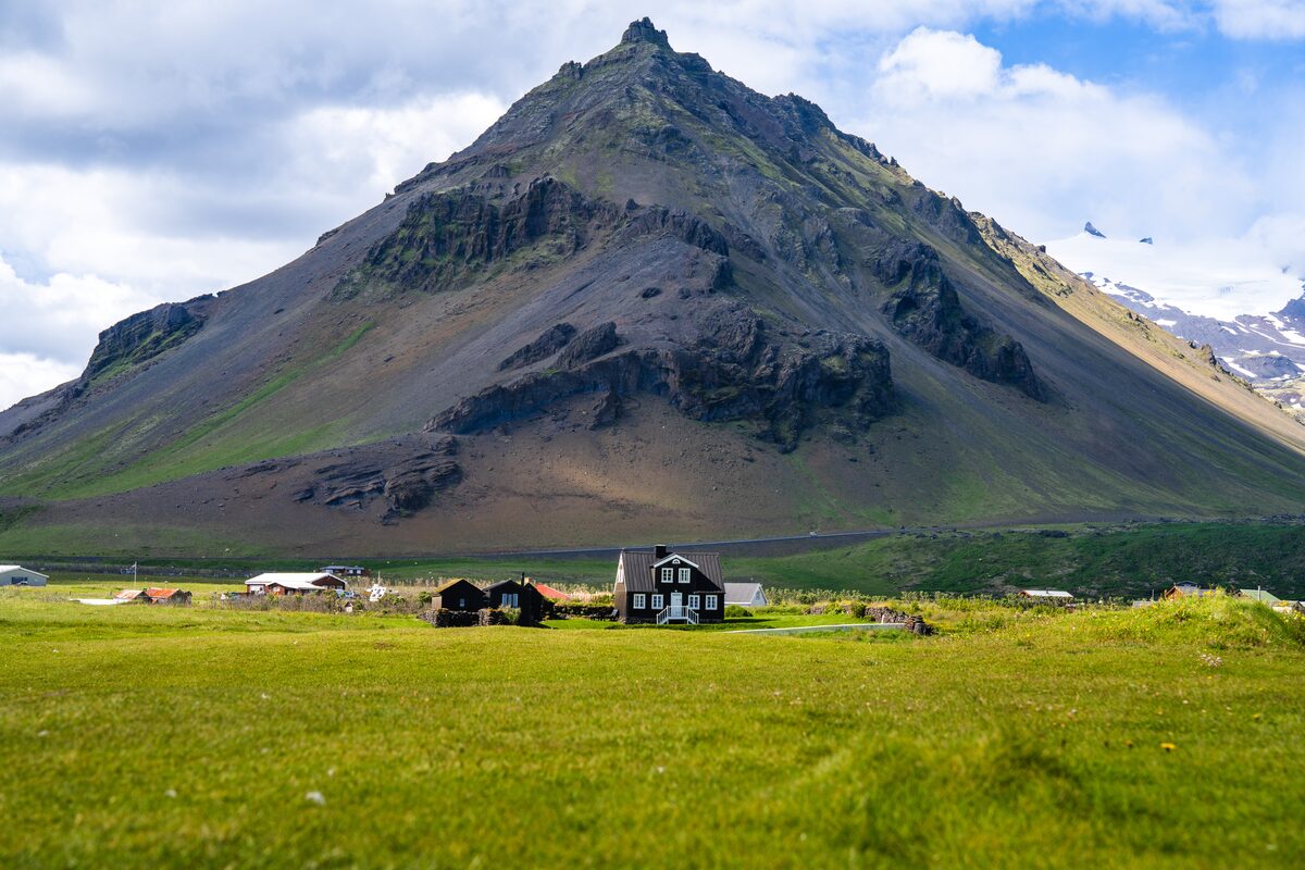 A dark timber house and turf-roofed buildings below the pyramid-shaped Stapafell at Arnarstapi.