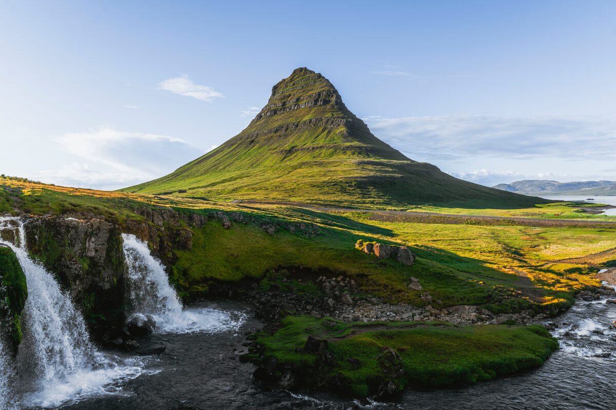 Kirkjufell's cone rising behind the tiered Kirkjufellsfoss waterfall and green riverbanks.