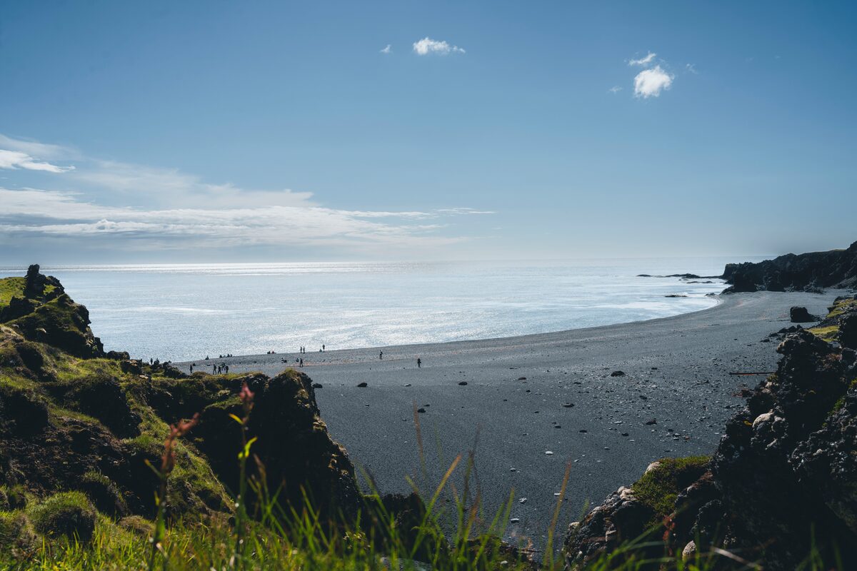 A black pebble beach curving toward dark cliffs, with tiny figures near the calm sea.