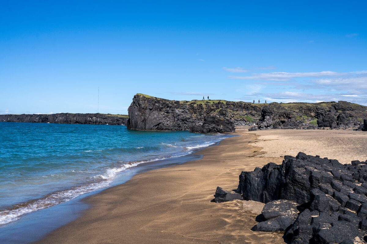 Golden sand and turquoise water at Skarðsvík, framed by black lava and a green-topped headland.