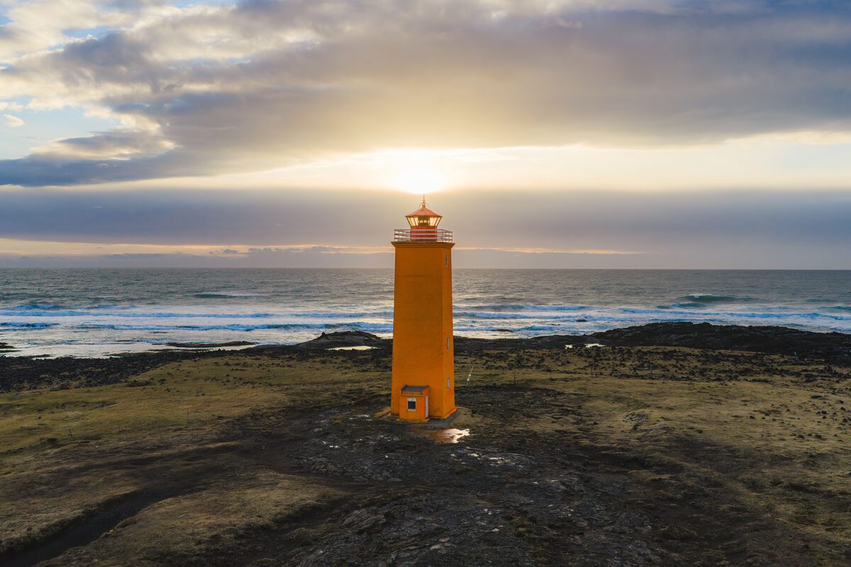 An orange lighthouse on a dark lava headland with the low sun behind it and waves beyond.