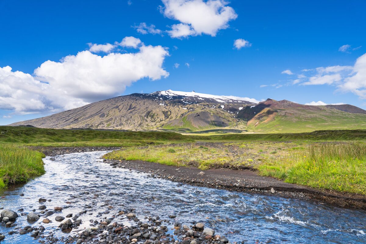 Snæfellsjökull's snow-capped summit beyond green banks and a shallow river with pebbles.