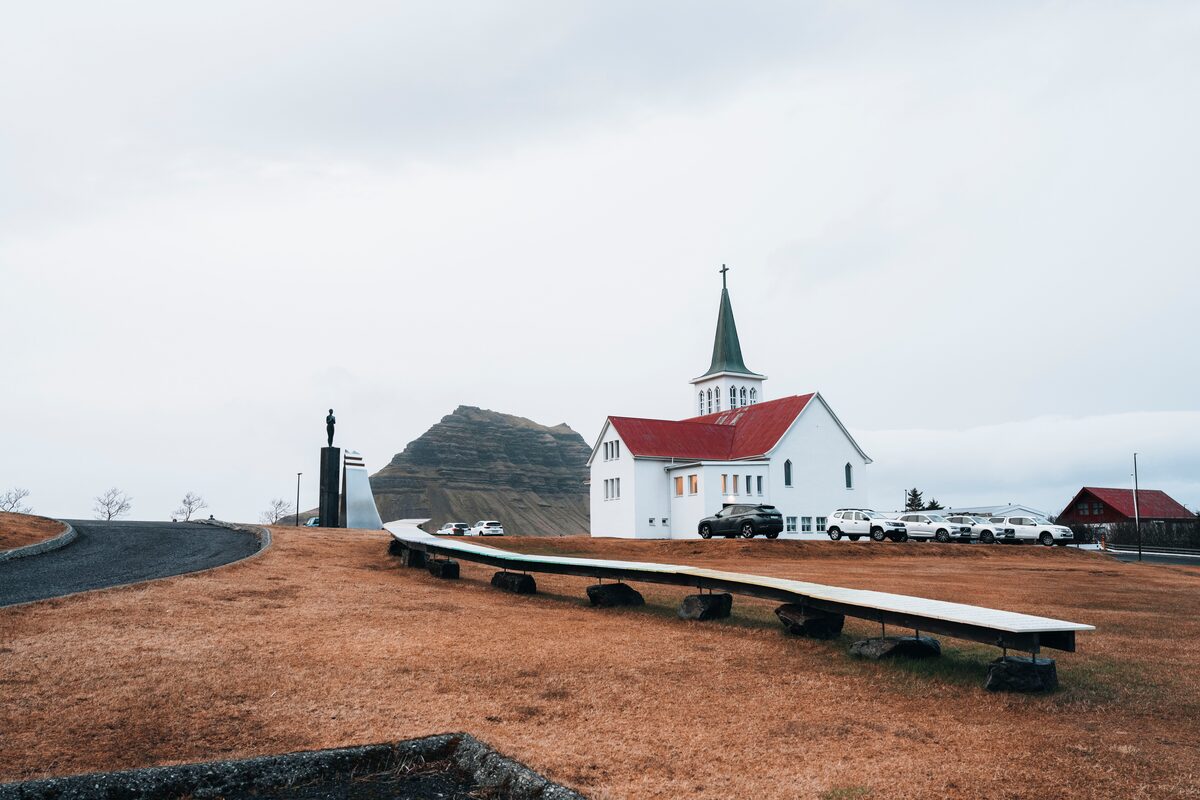 A white church with a red roof behind a long white curved structure on wooden supports.
