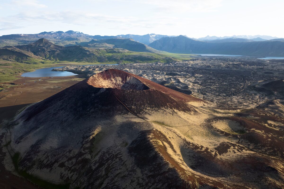 A red scoria crater with a hollow summit above a dark lava field, lake and mountains beyond.