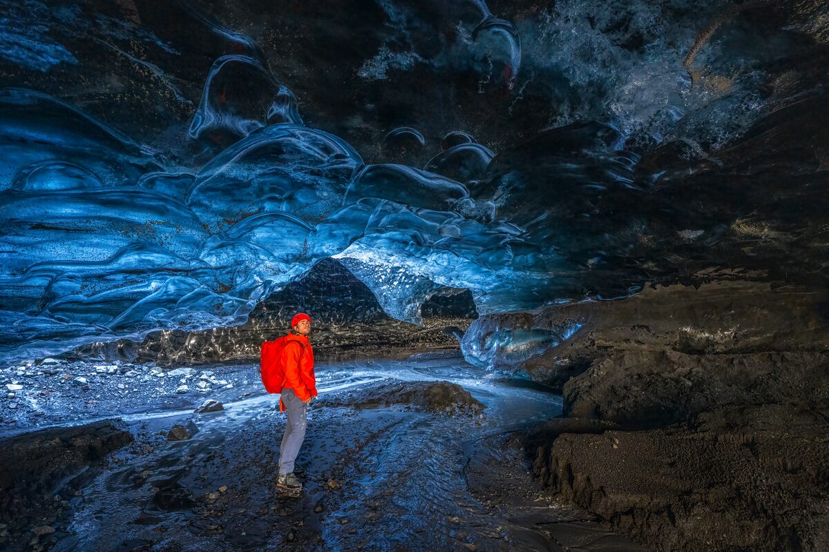 Male wearing red jacket and safety helmet with torch admiring ice cave walls inside crystal ice cave.
