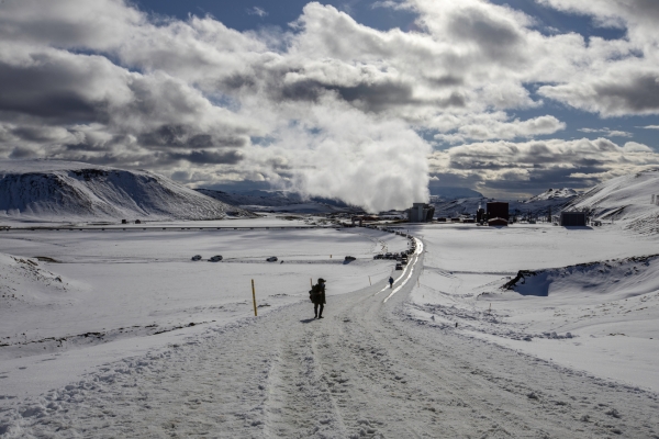 A person walks on a snow-covered road at Krafla toward the power station's steam plume.