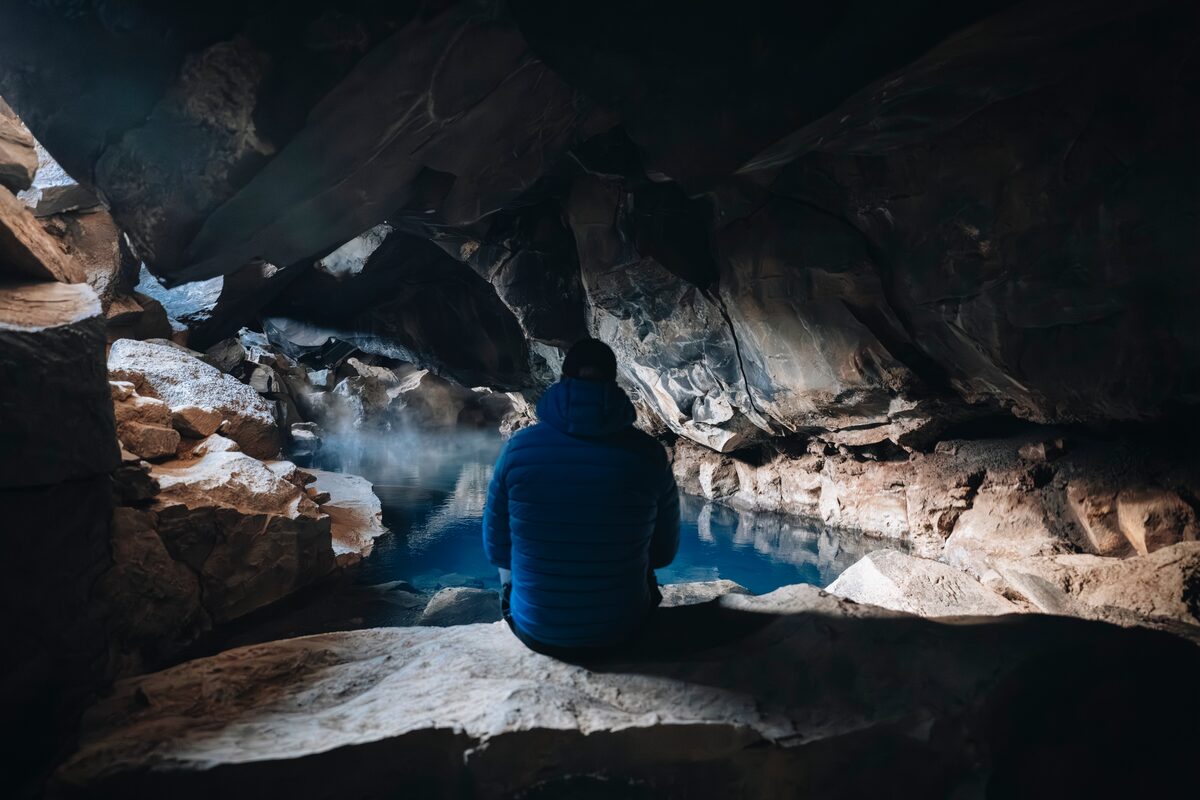 Person sitting inside Grjótagjá lava cave, looking at a glowing blue geothermal pool.