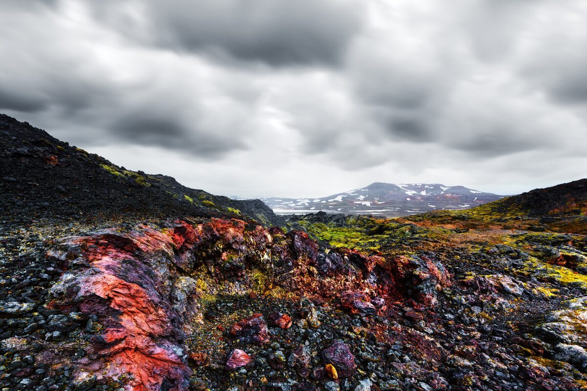 Red, orange, and black rock at Krafla lava field, moss growing over it, mountains behind.