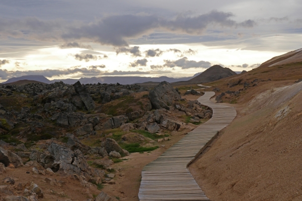 Wooden boardwalk winding through reddish volcanic terrain at Leirhnjúkur, hills in the distance.