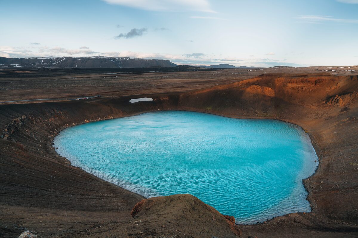 Turquoise crater lake inside Víti's dark volcanic rim, orange-tinted slopes, mountains in the distance.