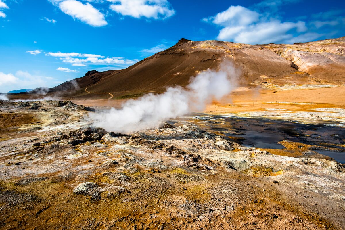 Steam rising from a fumarole in a sulfur-stained geothermal field, orange-toned hills behind.