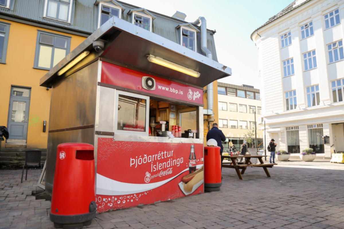 The red Bæjarins Beztu Pylsur hot dog kiosk on a downtown Reykjavík plaza, with picnic tables.