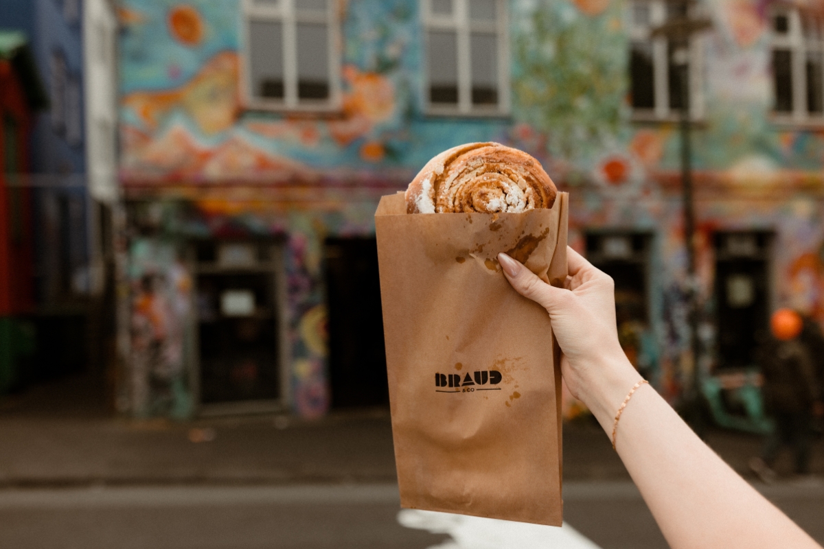 A hand holding a Brauð & Co paper bag with a cinnamon bun outside its mural-covered Reykjavík bakery. 