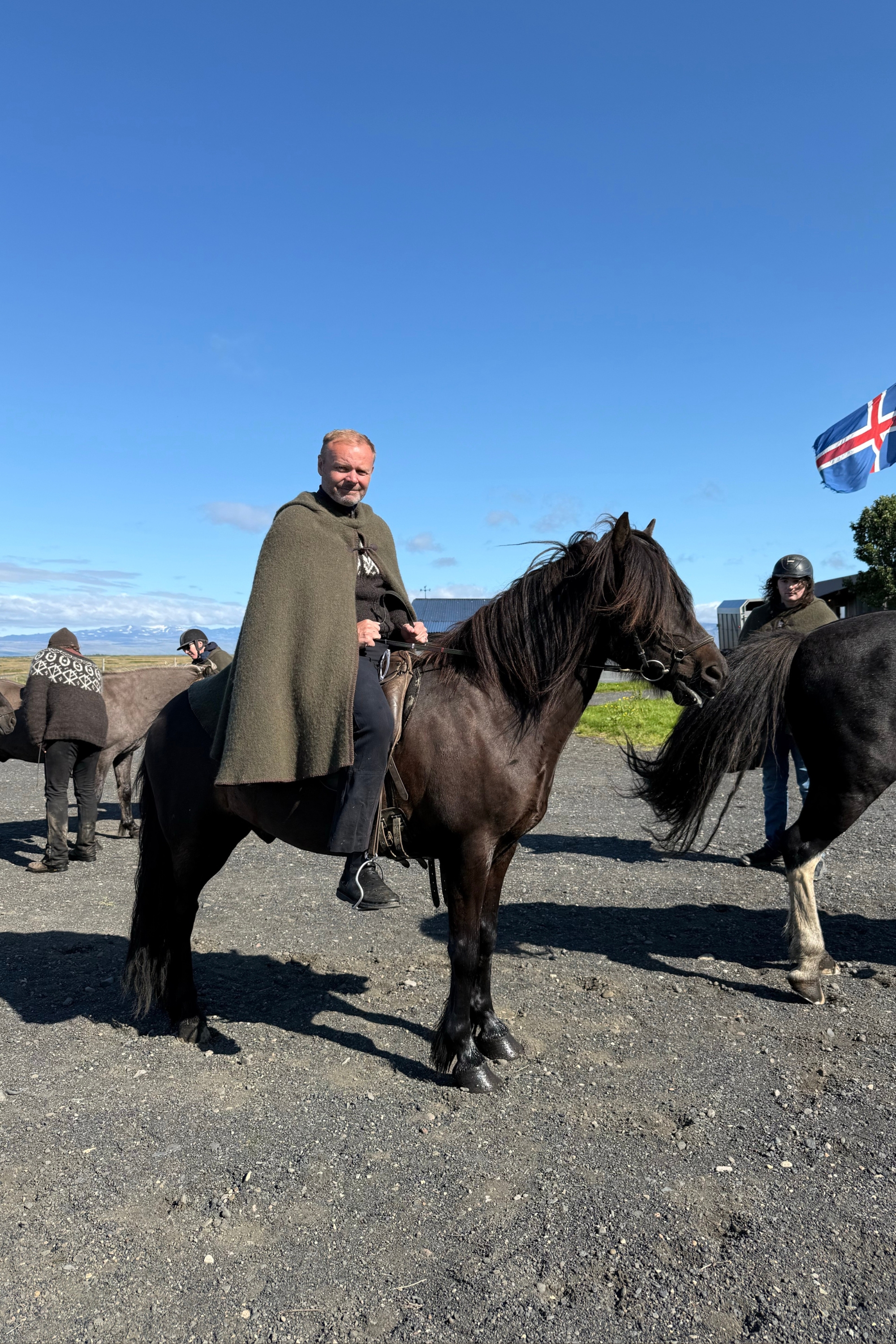 Man in a green cape standing beside a dark Icelandic horse under the Icelandic flag.