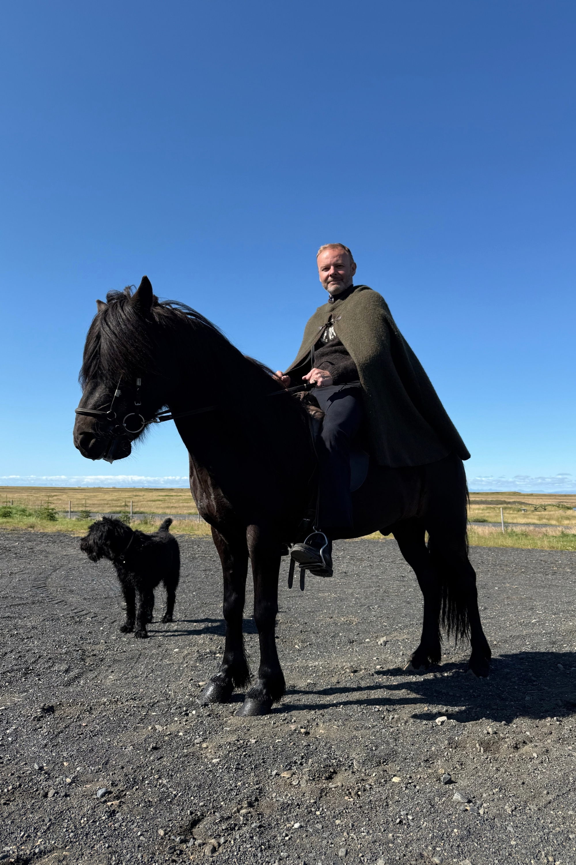 Man in a green cape mounted on a black Icelandic horse with a black dog standing nearby.