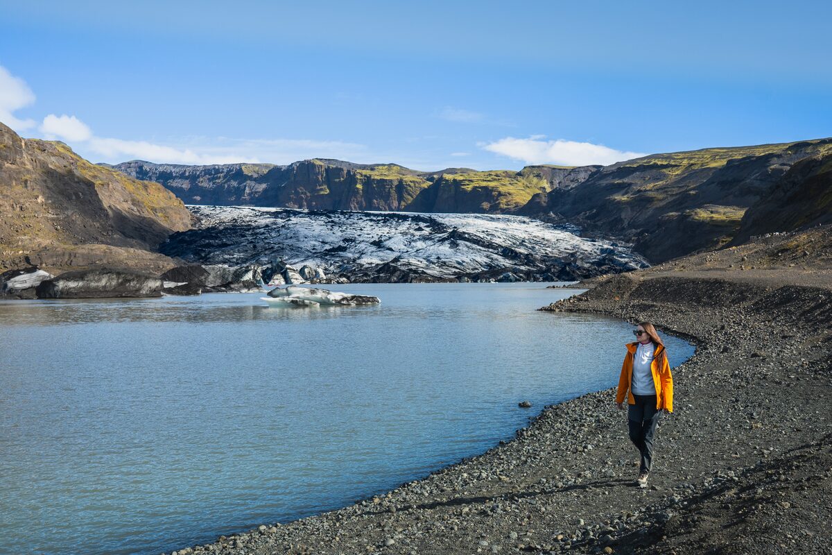 Female tourist walking on rocky ground next to Solheimajokull glacier river, views of Solheimajokull glacier in background.