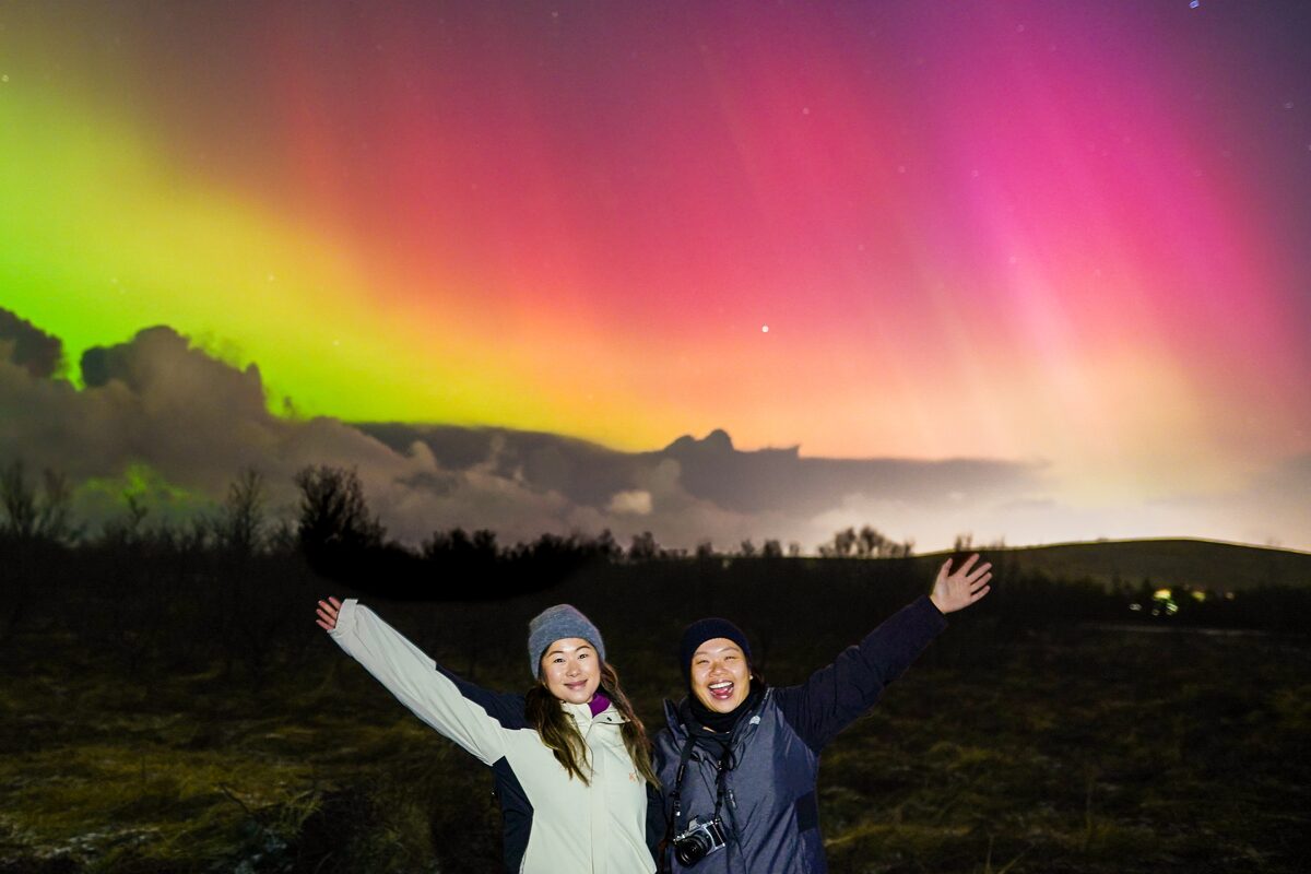 Two smiling people with raised arms under a pink and green aurora in Southwest Iceland’s countryside.