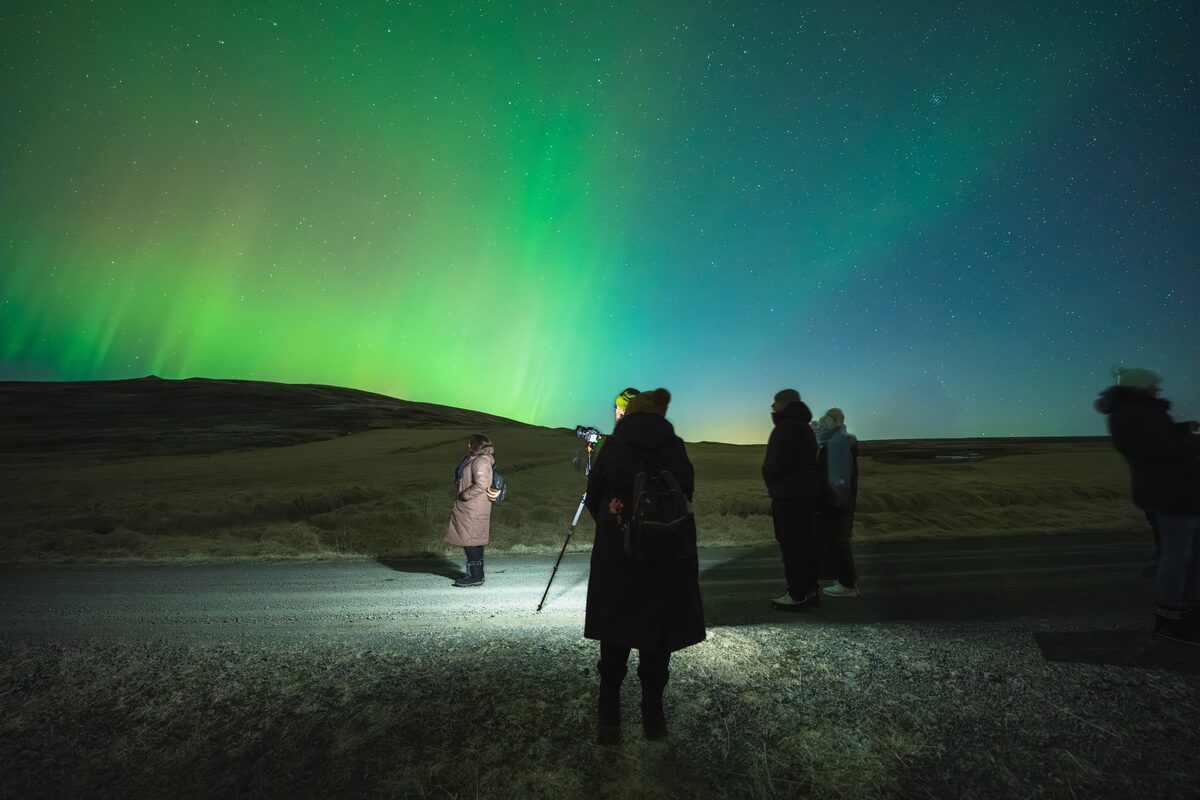 Group with a camera on a tripod under green northern lights beside a road in Southwest Iceland.