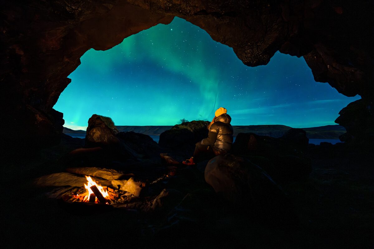 Person by a campfire looking out from a cave at a green aurora in Iceland.