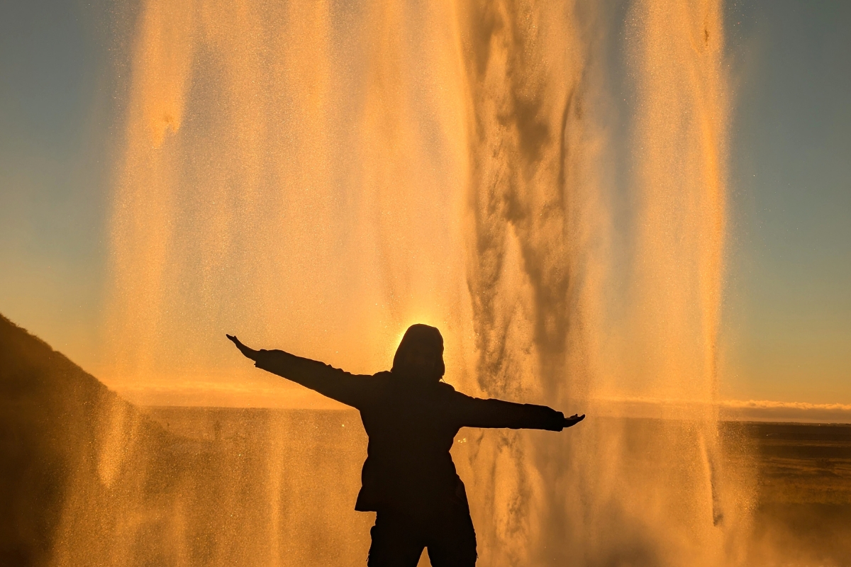 Neda silhouetted with arms outstretched behind Seljalandsfoss, the water lit gold by the sun.