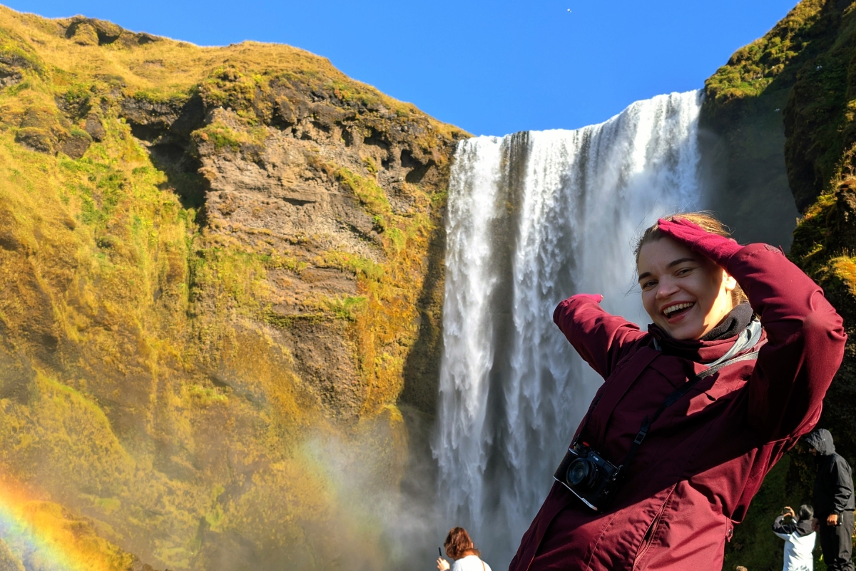 Neda laughing in front of Skógafoss, with a rainbow in the spray and other visitors below.