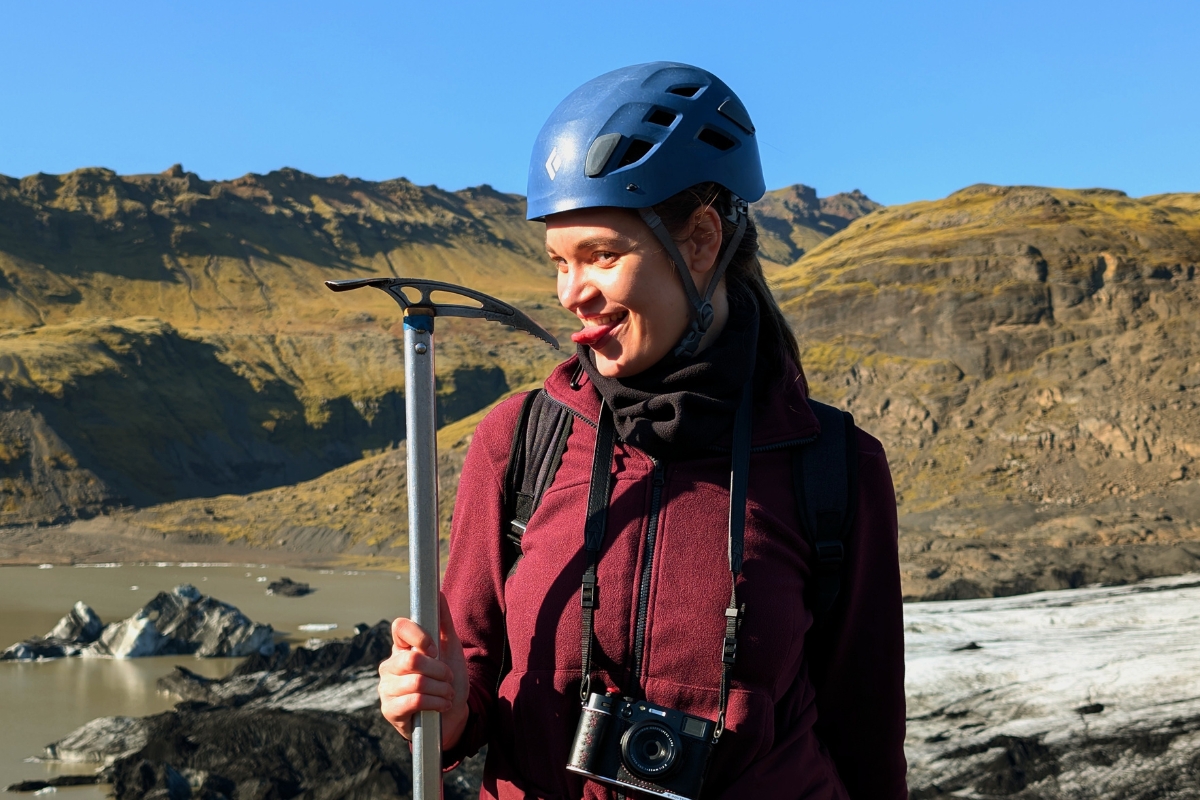 Neda grinning in a blue helmet with an ice axe on Sólheimajökull, the glacier lagoon behind her.