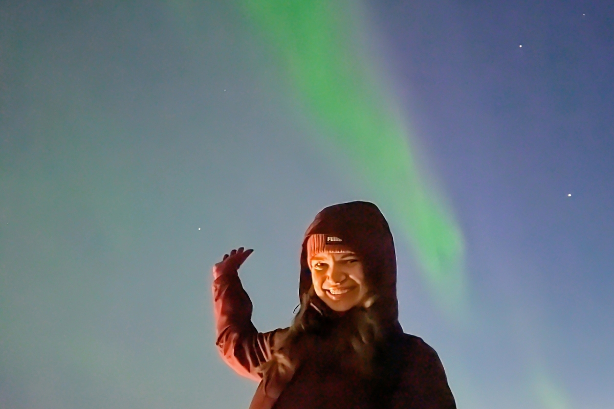 Neda waving under a green band of Northern Lights in the night sky over Reykjavík.