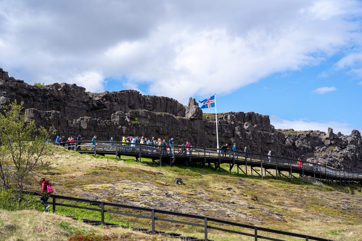 Visitors walk below rocky cliffs and an Icelandic flag at Þingvellir National Park.