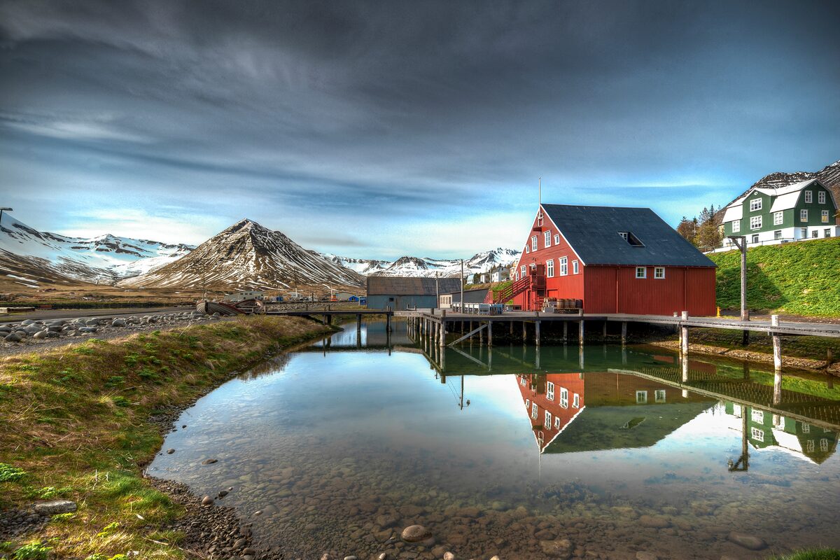 Red Herring Era Museum building on a pier reflected in calm water below snowy mountains.