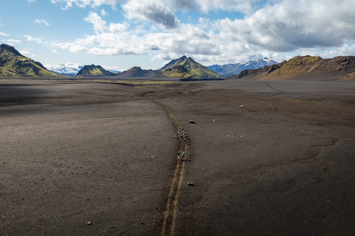 Hikers on a dirt road over a dark sandy plain toward green mountains on the Laugavegur trail.