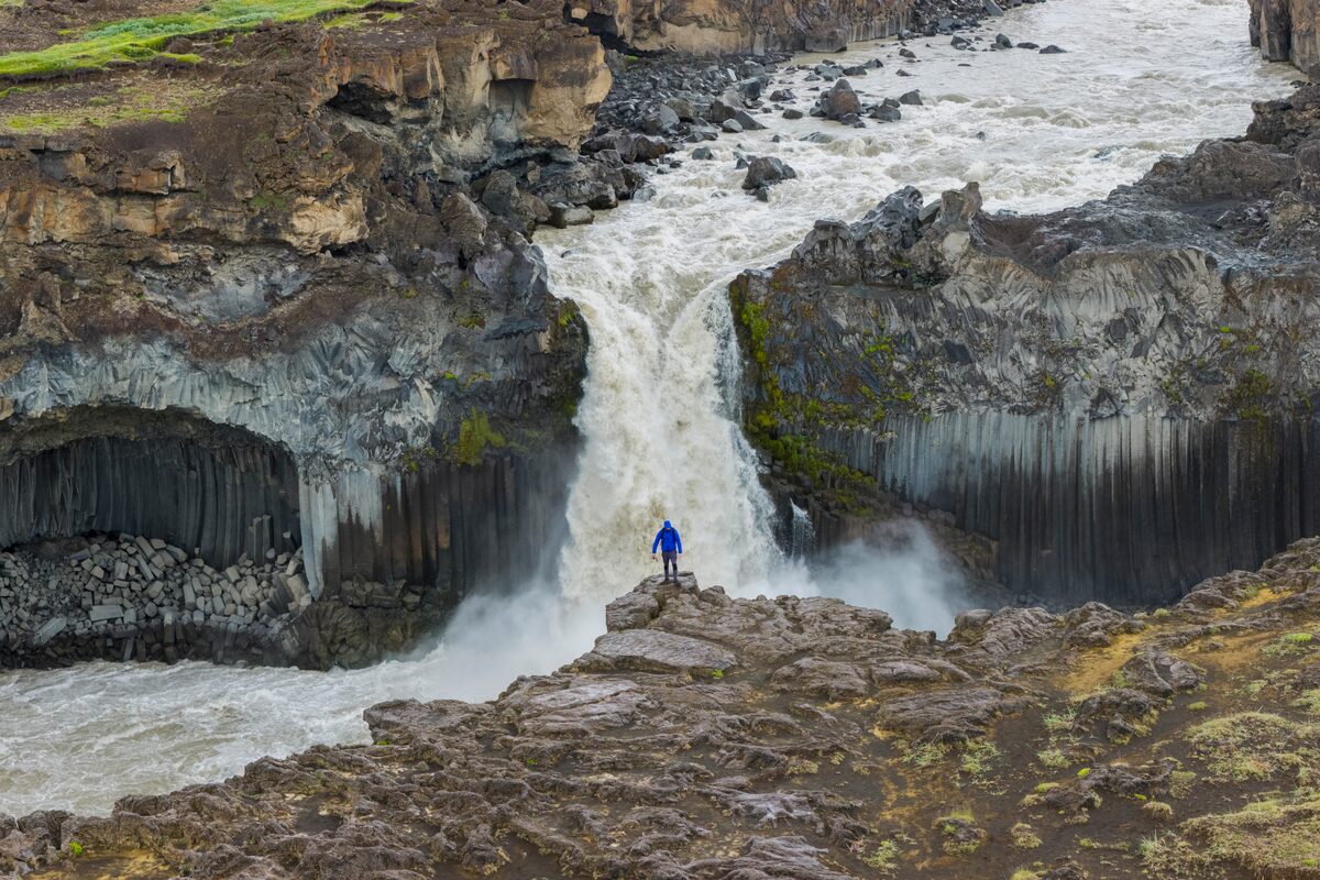 Person in a blue jacket on the rocks in front of Aldeyjarfoss waterfall and its black basalt columns.