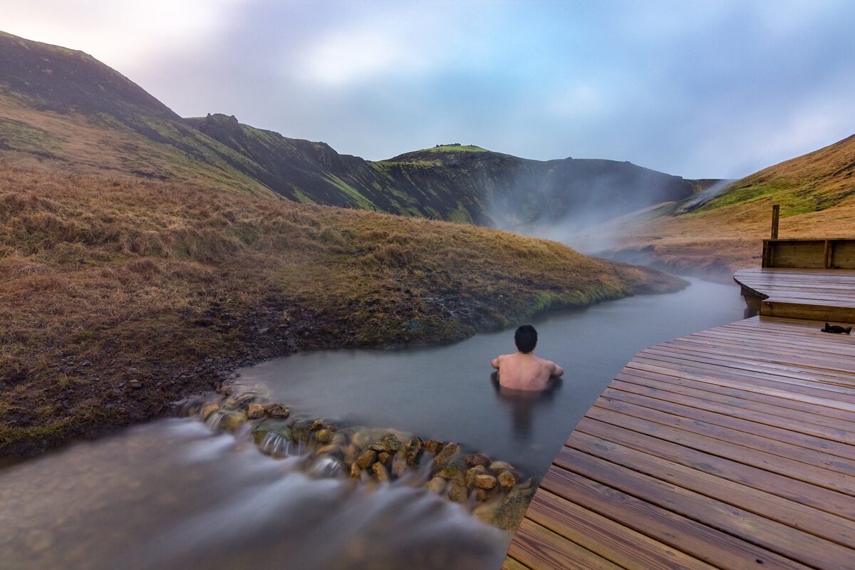 A person bathing in Reykjadalur’s steaming river beside a wooden boardwalk and grassy hills.