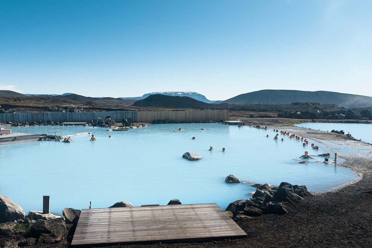 Bathers in the milky blue Earth Lagoon, with a wooden deck, dark rocks and hills beyond.