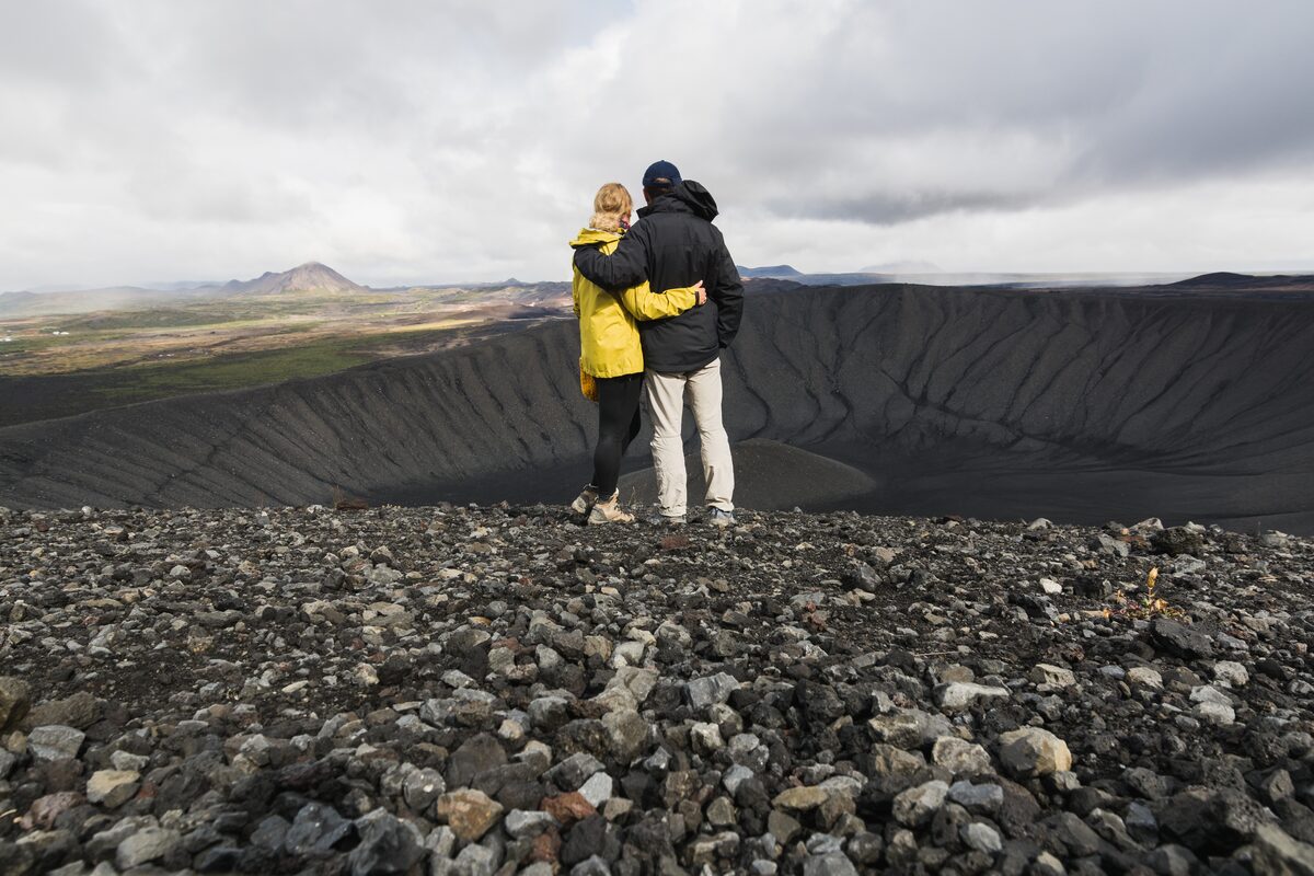 Two hikers on the rocky rim, arms around each other, looking into Hverfjall’s dark crater.