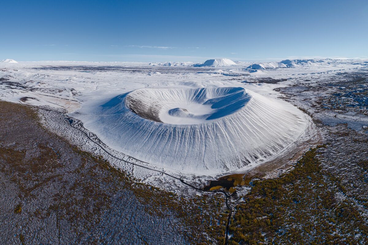 Aerial view of snow-covered Hverfjall crater on a snowy plain under a clear blue sky.