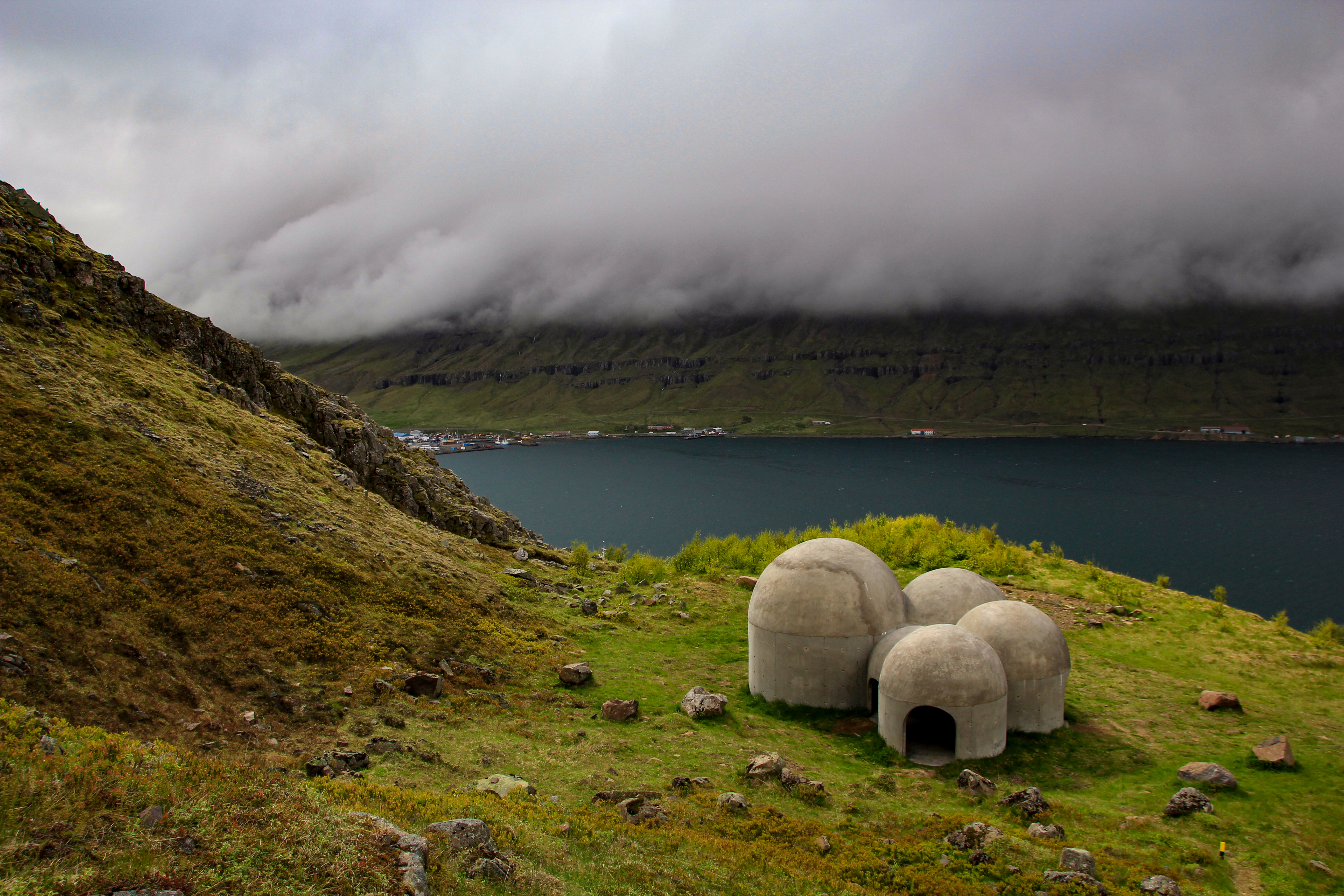 Tvísöngur’s concrete domes on a grassy hillside above a fjord under low cloud.